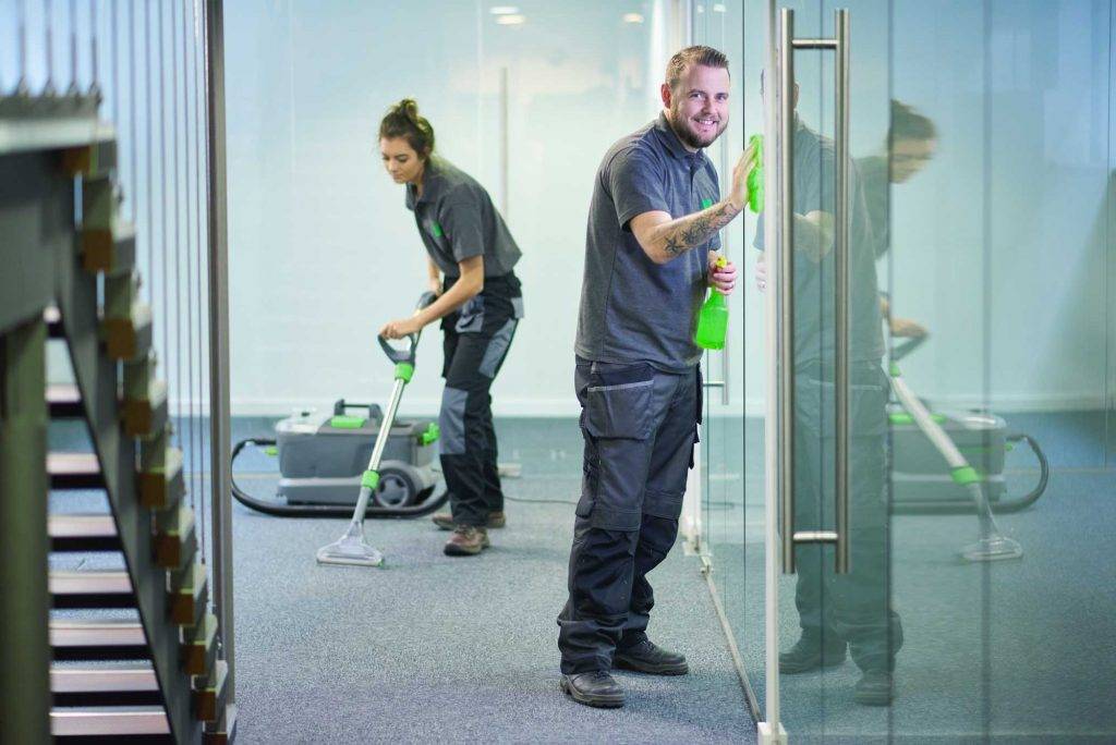 Zwei Mitarbeiter der Gebäudereinigungskonzepte Deutschland reinigen professionell ein Büro. Der eine Mitarbeiter wischt den Boden, während der andere die Glaswände mit einem Fensterreiniger säubert. Das Ergebnis: ein sauberes und gepflegtes Büro.